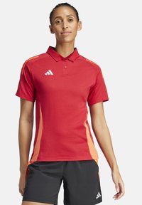 Rotes Polohemd mit orangefarbenen seitlichen Streifen, aus strukturiertem Stoff. Verfügt über einen Kragen und eine Knopfleiste mit drei Knöpfen. Adidas-Logo auf der Brust.