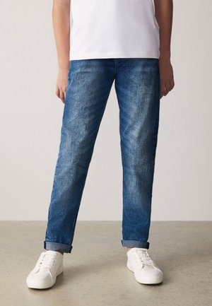 Personne portant un jean bleu retroussé, des baskets blanches et un t-shirt blanc, se tenant sur un sol lisse devant un fond uni.