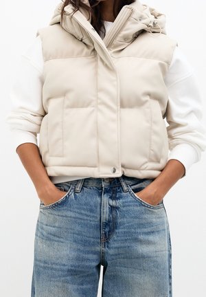 Veste matelassée beige sans manches avec poches portée sur un haut blanc à manches longues, associée à un jean bleu délavé. Mains dans les poches.