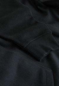 Schwarzer Hoodie mit gerippten Bündchen, weichem Material und einer Kängurutasche. Die Nähte sind ordentlich, und der Stoff wirkt dick und warm.