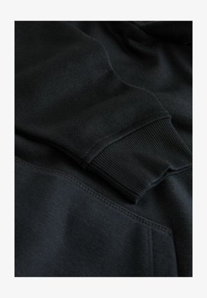 Hanorac negru cu mansete ribbed, textură moale și buzunar tip kangur. Cusăturile sunt îngrijite, iar materialul pare gros și călduros.