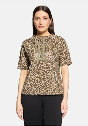 Vrouw met steil bruin haar draagt een bruin-beige korte mouw top met luipaardprint, voor met trekkoorden, en zwarte broek, staand stil.