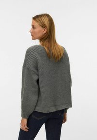 Szary, oversize'owy sweter z dzianiny o ribbowanej fakturze i obniżonych ramionach, z szerokim dekoltem i dłuższym tylnym zakończeniem. Noszony z ciemnymi dżinsami.