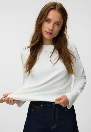 Jeune femme aux longs cheveux bruns portant une chemise blanche à manches longues et un jean bleu foncé, tirant légèrement la chemise vers l'avant.