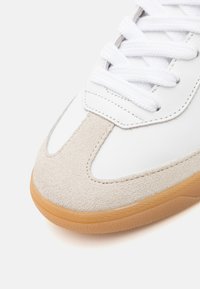 Adidași albi cu accente de suede bej și talpă din cauciuc caramel. Au șireturi albe plate și detalii cusute pe margini.