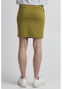 Jupe moulante vert olive avec une texture lisse, dotée de deux poches arrière et d'un ourlet juste au-dessus du genou. Assortie avec des baskets blanches.