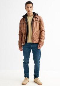 Chaqueta de cuero marrón con cremalleras y detalles cosidos, superpuesta a una sudadera negra. Combinada con jeans azules y zapatillas ligeras color tostado.