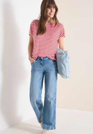 Kvinde smilende, iført rød- og hvidstribet skjorte, lyseblå vidde jeans, holder en sammenfoldet denimjakke i den ene hånd.