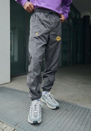 Osoba ubrana w szare spodnie Los Angeles Lakers, fioletową bluzę i białe buty sportowe Nike stoi przed budynkiem.