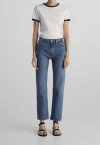 Person som bär en vit t-shirt med svarta detaljer, högmidjade blå raka jeans och svarta sandaler med öppen tå, stående mot en grå bakgrund.