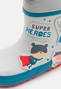 Botte de pluie en caoutchouc grise avec des accents bleus, ornée d'autocollants de super-héros en cartoon et d'une semelle rouge texturée. Les graphiques incluent des animaux et des étoiles.