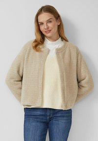 Beiger, strukturierter Cardigan mit rundem Ausschnitt und offenem Vorderteil, darüber ein hellcremiger Strickpullover, kombiniert mit blauen Jeans.
