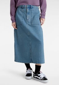 Falda de mezclilla, azul claro, larga con cintura alta y dos bolsillos frontales. Combinada con zapatillas negras y blancas. Diseño texturizado y casual.