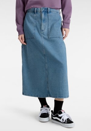 Falda de mezclilla, azul claro, larga con cintura alta y dos bolsillos frontales. Combinada con zapatillas negras y blancas. Diseño texturizado y casual.