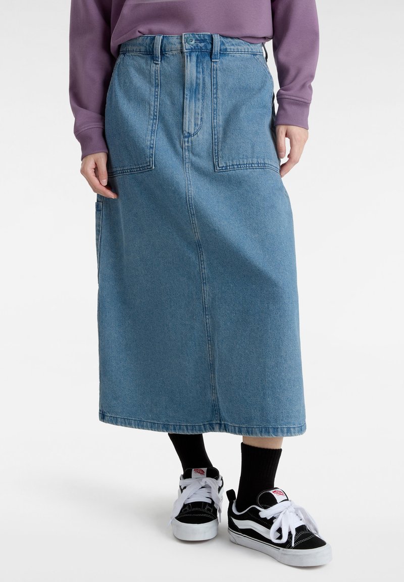 Falda de mezclilla, azul claro, larga con cintura alta y dos bolsillos frontales. Combinada con zapatillas negras y blancas. Diseño texturizado y casual.