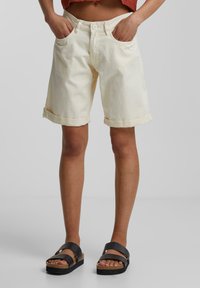 Shorts en denim crème clair avec ourlets retroussés, cinq poches et fermeture à bouton unique, accompagnés de sandales plates noires.