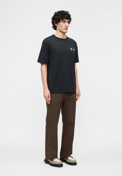 Άνδρας με σγουρά μαλλιά που φοράει μαύρο μπλουζάκι με δύο animal patches, καφέ παντελόνι και δίχρωμα παπούτσια, στέκεται μπροστά σε απλό φόντο.