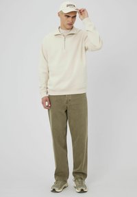 Sweatshirt beige à fermeture éclair avec un col haut, associé à un pantalon vert et des baskets beiges. Une casquette blanche avec le texte "GEORGETOWN".