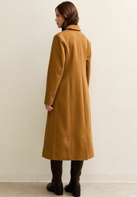 Vrouw die met haar hoofd naar links draait, gekleed in een lange camel kleurige jas en donkere bruin kniehoge laarzen, tegen een effen beige muur.