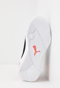 Semelle de sneaker blanche avec des motifs de grip texturés et un logo Puma rouge, positionnée debout contre un mur d'angle blanc.