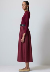 Vestido burdeos de manga larga con falda fruncida y cinturón negro. La modelo lleva zapatos planos beige, que destacan por un diseño elegante y minimalista.