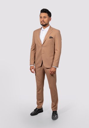 Mann in beigem Anzug mit weißem Hemd und Taschentuch, trägt schwarze Dressschuhe vor einem schlichten hellen Hintergrund.