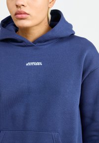 Sweatshirt azul-marinho feito de mistura de algodão, apresentando um bolso frontal e um logótipo em texto branco "JORDAN EST. 1984" bordado no peito.