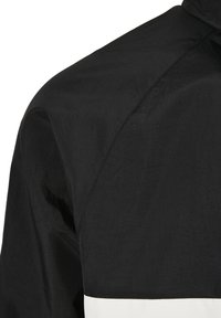 Giacca a vento nera con finitura testurizzata, caratterizzata da un pannello bianco nella parte inferiore e un colletto alto. Design pulito con hardware minimale.