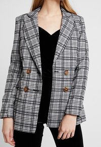 Dvojité sako v černobílém kostkovaném vzoru, s hnědými knoflíky, ostrými klopami a přesným střihem.