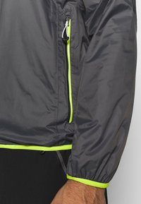 Veste grise légère avec des accents jaunes réfléchissants, des poignets élastiques et une poche zippée. Fabriquée en matériau lisse et résistant à l'eau.