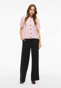 Svjetloružičasti kardigan s kratkim rukavima i velikim smeđim gumbima, uparен s crnim hlačama širokih nogavica. Glatka tekstura, opušteni kroj, ležerni dizajn.