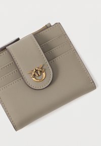 Carteira de couro cinza com fecho de aba e emblema de pássaro em tom dourado. Apresenta três divisórias para cartões na parte da frente e uma superfície texturizada.