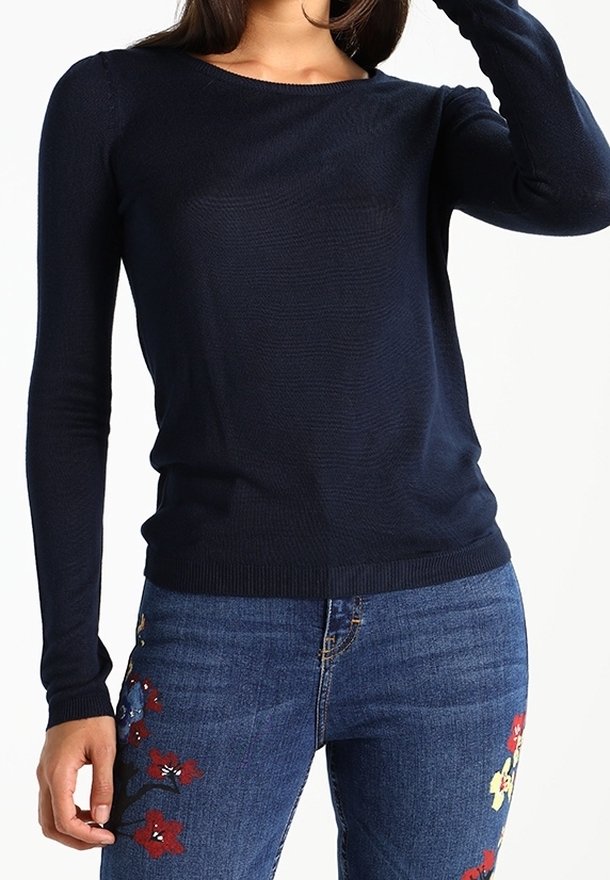 Femme portant un pull ajusté bleu marine à manches longues et un jean bleu brodé de fleurs rouges et jaunes sur les cuisses.