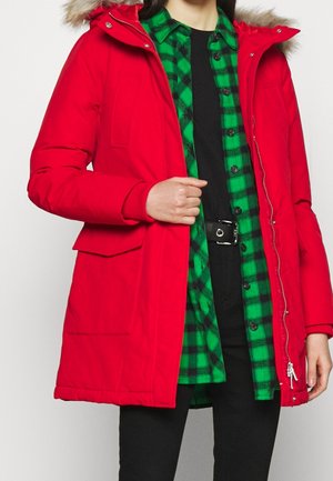 Red puffer jacket with a fur-lined hood, featuring a front zipper, side pockets, and ribbed cuffs, layered over a black and green plaid shirt.