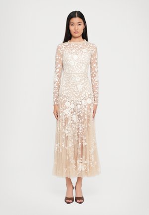 Mujer vestida con un vestido largo, de color nude, transparente, con bordado floral blanco y mangas largas, de pie contra un fondo blanco liso.