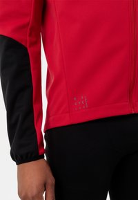 Veste rouge avec manches noires, fabriquée en matériau lisse. Dispose d'une fermeture éclair, de couleurs contrastantes et d'un texte imprimé à l'ourlet.