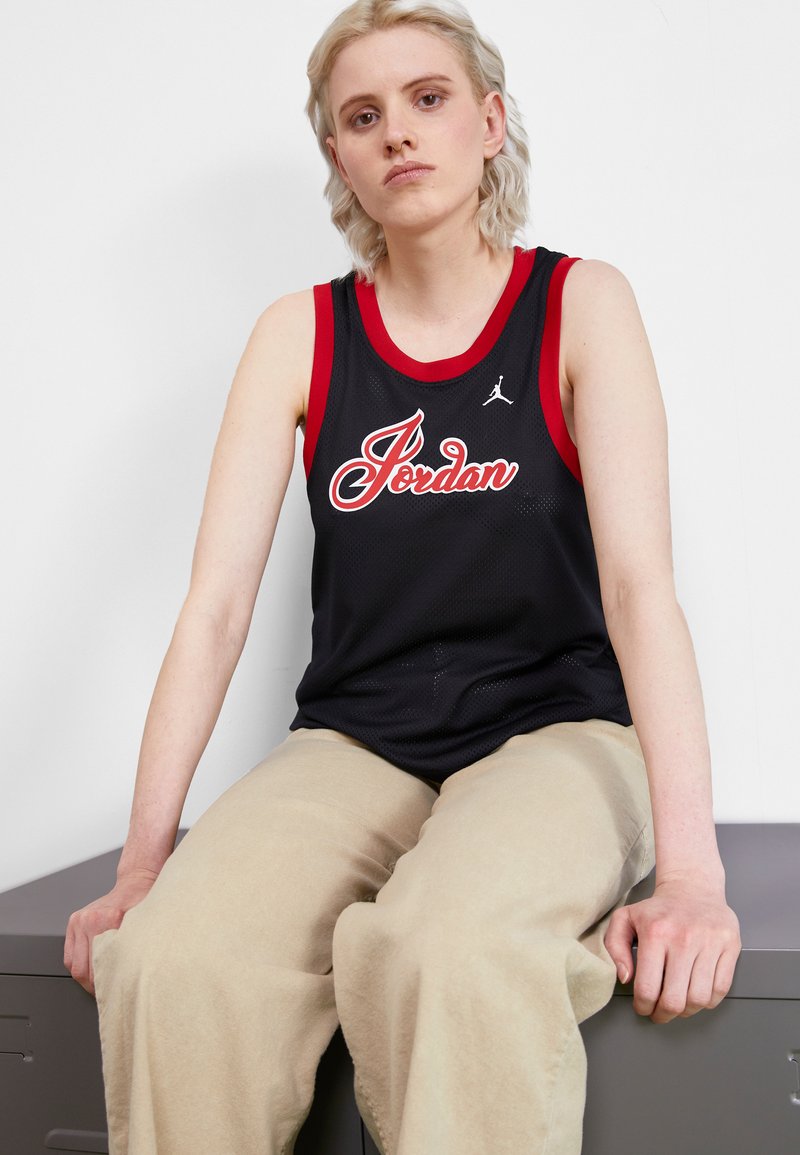 Black basketball jersey with red accents, featuring "Jordan" in white script and a small Jumpman logo. Model sits on a gray surface, wearing beige pants.