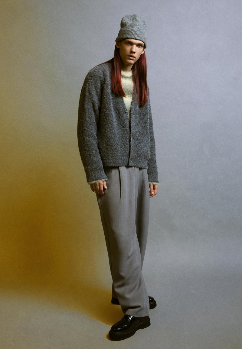Cardigan grigio con trama, chiuso con bottoni, indossato sopra un maglione crema, abbinato a pantaloni grigi a vita alta e scarpe con plateau nere.
