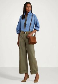 Sieviete ar zilā pogātā krekliņa, ar rakstainu topu, zaļajām platajām biksēm, smilškrāsas papēžiem un brūnu plecu somu.