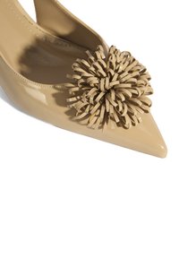 Chaussure en cuir verni beige à bout pointu, ornée d'un accent décoratif en forme de fleur texturée faite de bandes torsadées dans une teinte similaire.