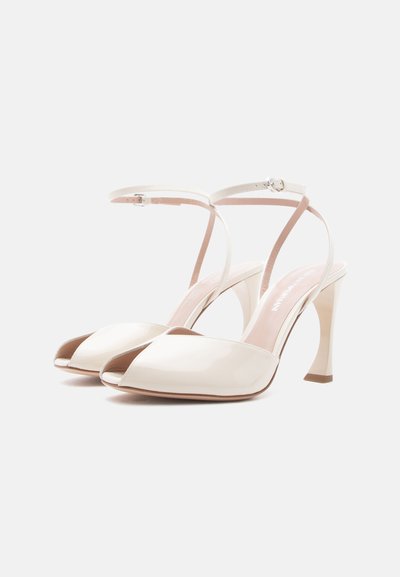 Πέδιλα από πατινέ δέρμα σε κρεμ απόχρωση με ανοιχτά δάχτυλα, λουριά στον αστράγαλο και ασημένια αγκράφα, σε λευκό φόντο.