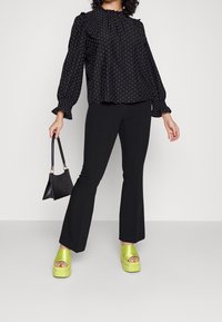 Zwarte blouse met stippen en pofmouwen, gepaard met zwarte flared broek en limoen groene plateausandalen. Houdt een kleine zwarte handtas vast.
