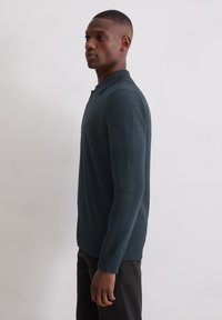 Ciemnoturkusowy sweter dziany o fakturowanym wykończeniu, z długimi rękawami i kołnierzem. Wykończenia z prążkowanego ściągacza na dole i mankietach. Noszony z czarnymi spodniami.