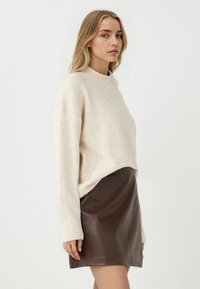 Maglione lavorato a maglia beige chiaro con collo alto e polsini a costine, abbinato a una minigonna in pelle marrone lucida. Texture liscia.