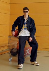 Džínová bunda, bílá košile s modrým logem, černé kalhoty a hnědé tenisky. Sedící s basketbalem vedle vozíku s míči.