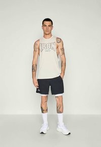 Uomo tatuato che indossa una maglietta senza maniche bianca con la scritta "IRON", pantaloni corti neri, calzini bianchi e sneaker bianche, in piedi su uno sfondo neutro.