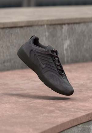 Zapatilla deportiva de color gris oscuro con parte superior de malla y ante, detalles texturizados, puntera redonda y cordones planos. Cuenta con una suela de goma flexible.