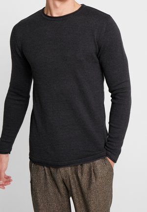 Homme portant un pull en maille noir ajusté à manches longues et un pantalon marron à motif chevrons, debout avec une main dans la poche.