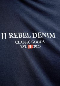 Mornarsko modro blago z belim besedilom "JJ REBEL DENIM CLASSIC GOODS EST. 2023" in majhnim rdečim kvadratnim logotipom z belo črko "R".