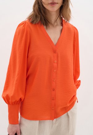 Vrouw draagt een feloranje V-hals blouse met lange gebolde mouwen en knoopsluiting, gecombineerd met lichtgekleurde broek.